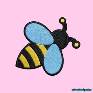 May include: An embroidered patch shaped like a bee with a black and yellow striped abdomen, a black head, two light blue wings, and two antennae with yellow tips. The design features a thick black border outlining the entire insect shape.