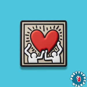 May include: Square patch featuring a red heart held by two white figures with raised arms. The design is set against a beige background with a black border. The patch has a black border and is on a blue background.