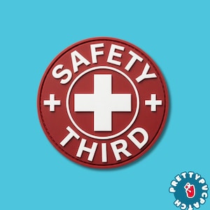 May include: A circular red patch featuring a white cross in the center. The word SAFETY is printed in white capital letters along the top curve, and the word THIRD is printed in white capital letters along the bottom curve, with small plus signs on each side.
