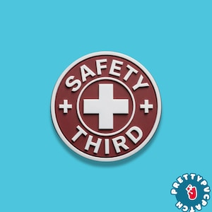 May include: A round, maroon patch with the words "SAFETY THIRD" in white lettering. A white cross is centered within the circle, with a white plus sign on each side. The patch has a white border and is set against a blue background.