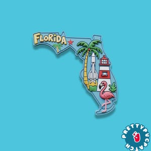 May include: A Florida-shaped magnet with a light blue outline, featuring a palm tree, rocket, lighthouse, and a pink flamingo. The word "FLORIDA" is written in yellow letters. The background is a solid light blue.