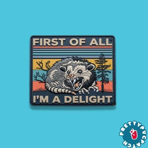 May include: A rectangular patch with a gray opossum illustration, open-mouthed and snarling. The patch has the text "FIRST OF ALL" above and "I'M A DELIGHT" below, with a retro sunset design in the background.