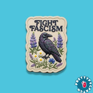 May include: A rectangular patch with the words "FIGHT FASCISM" above a detailed illustration of a raven, surrounded by flowers. The raven is black with iridescent purple and green feathers. The patch has a cream-colored border and is set against a blue background.