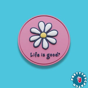 May include: A round, pink coaster with a white and blue daisy design in the center. The text "Life is good!" is printed below the flower. The coaster has a light blue background.