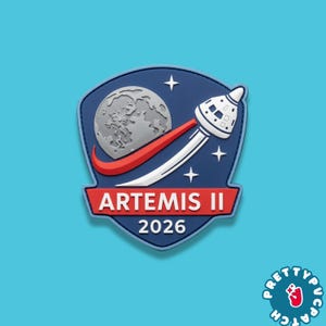 May include: A blue, red, and white Artemis II patch featuring a stylized rocket launching towards a gray moon. The patch includes the text "ARTEMIS II" and "2026" in white lettering on a red banner.