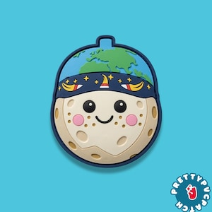 May include: A whimsical, cartoon-style patch featuring a smiling moon face with rosy cheeks and a blue headband adorned with stars and bananas. The top of the patch depicts the Earth. The patch is set against a bright blue background.