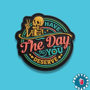 May include: A circular black rubber patch featuring a yellow skeleton making a peace sign. The text reads HAVE THE DAY YOU DESERVE in pink, teal, and yellow lettering. The design includes concentric circles in green, teal, and yellow colors.