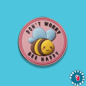 May include: A round, pink patch with a cartoon bee and the words "DON'T WORRY BEE HAPPY" in black. The bee is yellow and brown with blue wings. The patch has a black border and is set against a blue background.