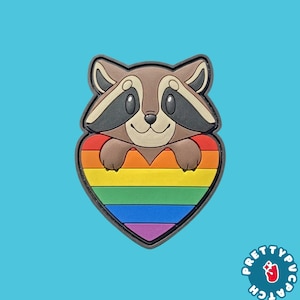May include: A rubber patch featuring a brown raccoon with black eye markings peeking out from behind a heart-shaped rainbow graphic. The design includes horizontal stripes of red, orange, yellow, green, blue, and purple. The text PRETTYPUCPATCH is visible.