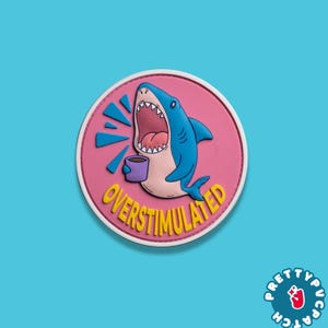 May include: A round patch featuring a cartoon shark with an open mouth, holding a coffee cup. The shark is blue, with a pink mouth and a white belly. The patch has the word "OVERSTIMULATED" in yellow text.