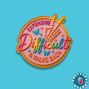 May include: A round, pink patch with the words "Expensive, Difficult, & Talks Back" in colorful, bubble letters. A skeleton hand makes a peace sign. Butterflies and stars decorate the patch. The patch has a gold border.