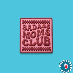 May include: A pink patch with the words "BADASS MOMS CLUB" in a bold, dark red font. The patch has a stitched border and a light pink background. The patch is on a blue background.