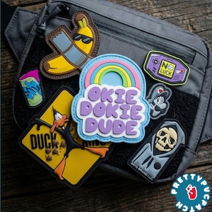 May include: A gray tactical fanny pack with several embroidered patches. Patches include a banana wearing sunglasses, a rainbow with the words "Okie Dokie Dude", and a cartoon duck. Other patches feature a video game controller and a grim reaper.