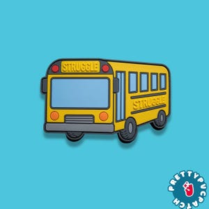 May include: A yellow school bus-shaped charm with the word "STRUGGLE" displayed on the front and top. The charm has black tires, a blue windshield, and red and orange accents. The background is a solid teal color.