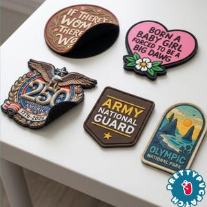 May include: A collection of colorful patches with various designs and text. One patch is a brown oval with the text "IF THERE'S WOM THERE WA". Another is a pink heart with the text "BORN A BABY GIRL FORCED TO BE A BIG DAWG". Other patches include military and patriotic themes.