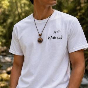 May include: White t-shirt with the word "Nomad" and a footprint graphic. A brown necklace with a wooden pendant featuring a mountain scene is worn. The shirt is short-sleeved and made of a soft material.