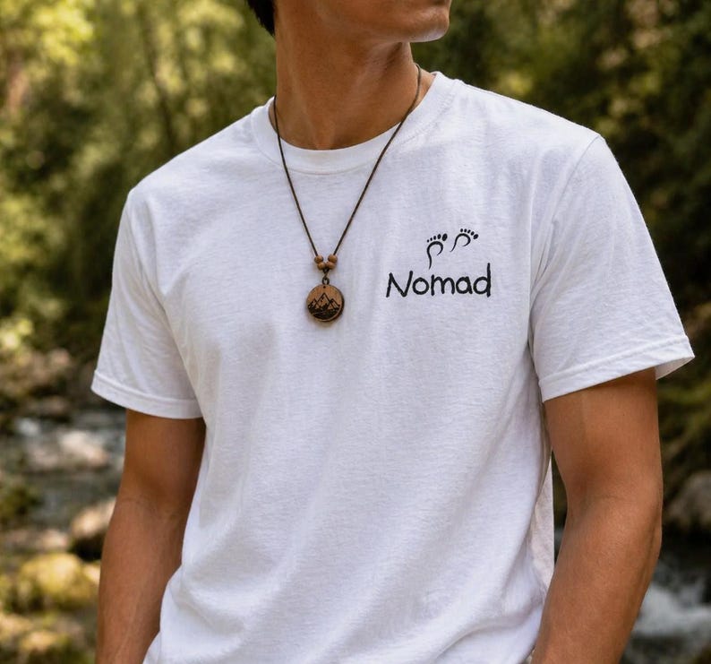 May include: White t-shirt with the word "Nomad" and a footprint graphic. A brown necklace with a wooden pendant featuring a mountain scene is worn. The shirt is short-sleeved and made of a soft material.