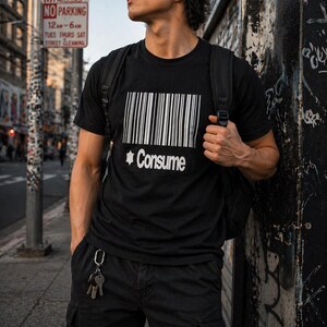 Puede incluir: Camiseta negra de cuello redondo con un diseño de código de barras blanco y la palabra "Consume" impresa debajo. La camiseta la lleva una persona con una mochila negra y pantalones cargo negros. Las llaves cuelgan del bolsillo.