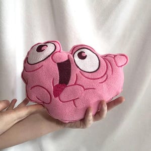 Op de afbeelding: Een roze pluche speelgoed in de vorm van een cartoongezicht, met grote ogen en een open mond. Het speelgoed heeft geborduurde details in donkerder roze en bruin. Het wordt in de handen van een persoon gehouden.