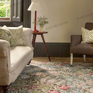 May include: A living room scene featuring a floral patterned rug with pink, green, and blue hues. A cream-colored sofa with patterned pillows sits near a wooden side table with a red lamp. A brown armchair is also visible.