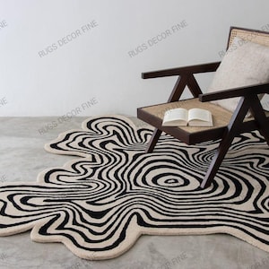 Könnte beinhalten: Abstrakter Teppich mit einem wellenförmigen Muster in Schwarz und Creme. Er liegt auf einem Betonboden, neben einem dunklen Holzstuhl und einem Buch. Der Stuhl hat eine gewebte Sitzfläche und Rückenlehne sowie ein cremefarbenes Kissen. Das Muster des Teppichs ähnelt einem flüssigen Design.