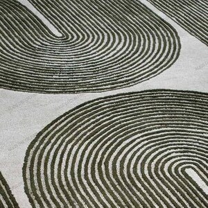May include: Close-up of a rug featuring a repeating pattern of olive green curved lines set against a light grey backdrop. The lines create a textured, layered effect, suggesting a modern, abstract design. The rug's design is suitable for various interior styles.