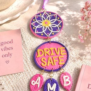 May include: A decorative car charm with a purple ribbon. The charm features a floral design, the words "DRIVE SAFE", and leaf-shaped pendants with letters. The color scheme includes pink, purple, yellow, and white.