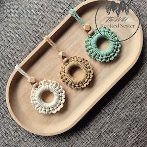 May include: Three macrame ornaments in white, beige, and mint green, each with a circular design and a wooden bead accent. The ornaments are displayed on a wooden tray. The Wild Knotted Nester logo is visible.