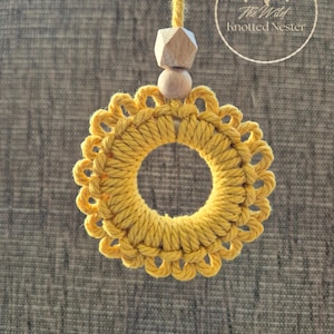 May include: A circular macrame ornament in mustard yellow featuring a looped edge design. The ornament hangs from a mustard yellow cord accented with a small wooden sphere and a faceted wooden bead. The text The Wild Knotted Nester is visible.