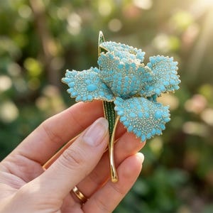 May include: A turquoise flower brooch with gold accents. The flower petals are covered in small, round turquoise stones, and the edges are trimmed with gold. The stem is gold with a green leaf. The brooch is held in a hand.