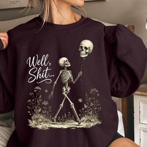 May include: A dark maroon crewneck sweatshirt featuring a graphic print of a skeleton walking through a field of flowers while holding a skull on a string like a balloon. The text "Well, Shit..." is printed in white script to the left of the skeleton.