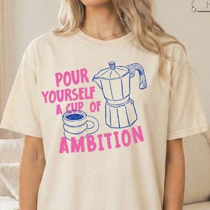 May include: An ivory short-sleeve t-shirt featuring a blue line-art illustration of a moka pot and a coffee mug. The text printed in pink reads, "POUR YOURSELF A CUP OF AMBITION." The shirt has a crew neckline and a relaxed, loose-fitting silhouette.