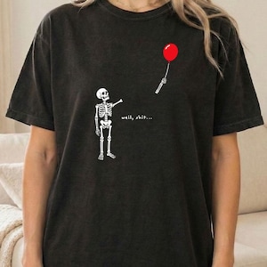 May include: A black short-sleeve t-shirt featuring a white graphic of a skeleton reaching for a floating red balloon. The text "well, shit..." is printed in white near the skeleton. The shirt has a crew neckline and a relaxed, loose fit.