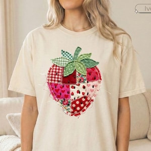 May include: Ivory-colored t-shirt featuring a large, patchwork strawberry design. The strawberry is composed of various fabric patterns, including gingham, hearts, and floral prints. The shirt has short sleeves and a relaxed fit, suitable for casual wear.