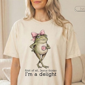 May include: An ivory-colored short-sleeve t-shirt featuring a graphic of a green frog wearing a pink bow and holding a small pink mug. Below the frog, the text reads: first of all, Jesus thinks I'm a delight. The shirt has a crew neckline.