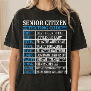 May include: Black t-shirt with the text "SENIOR CITIZEN TEXTING CODE" and a list of acronyms and their meanings, such as "BFF: BEST FRIEND FELL" and "LOL: LITTLE OLD LADY". The shirt is worn by a person.