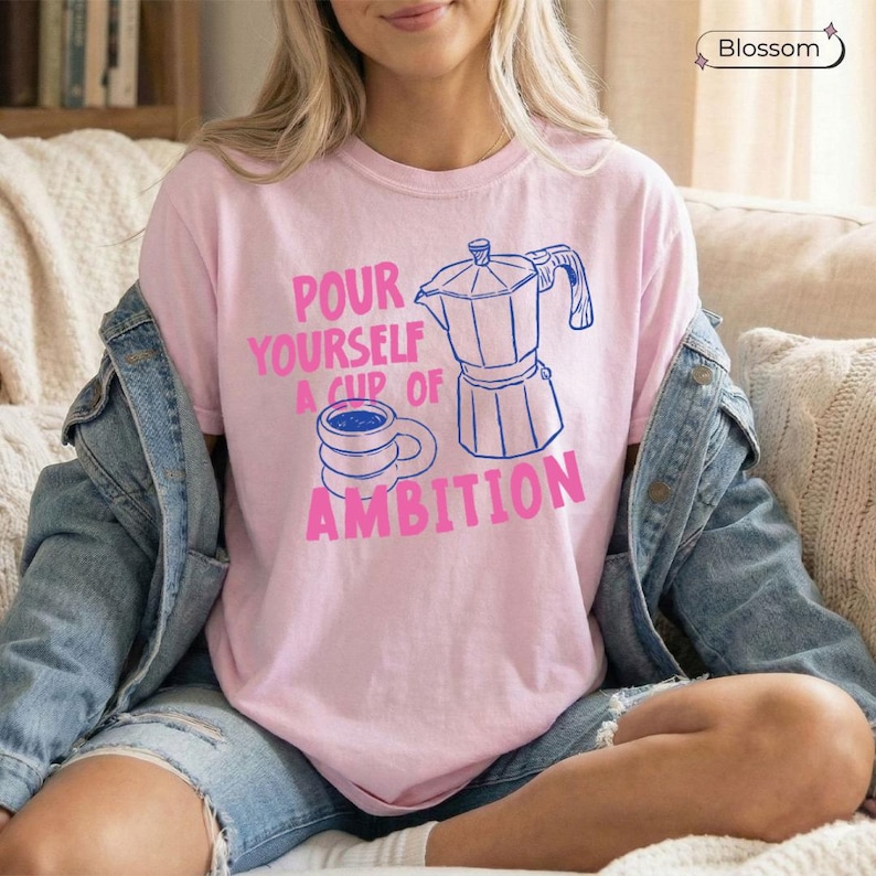 May include: A light pink short-sleeve t-shirt featuring a blue line-art graphic of a moka pot and a coffee mug. The text "POUR YOURSELF A CUP OF AMBITION" is printed in pink lettering. The shirt is worn under a blue denim jacket with blue denim shorts.