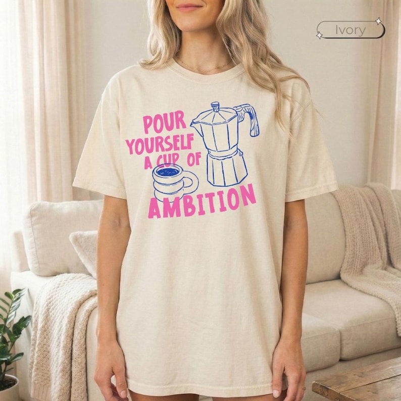 May include: An ivory short-sleeve t-shirt featuring a blue line-art illustration of a moka pot and a coffee mug. The text printed in pink reads, "POUR YOURSELF A CUP OF AMBITION." The shirt has a crew neckline and a relaxed, loose-fitting silhouette.