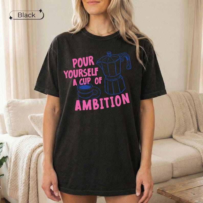 May include: A black short-sleeve t-shirt featuring a pink graphic print that reads "POUR YOURSELF A CUP OF AMBITION" alongside a blue outline illustration of a moka pot and a coffee mug. The shirt has a crew neckline and a relaxed fit.