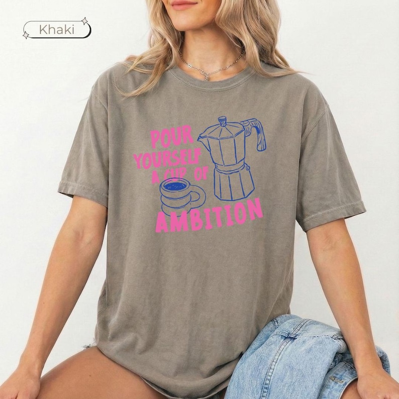 May include: A khaki short-sleeve t-shirt featuring a graphic print of a blue moka pot and a blue coffee cup. The text in pink reads, "POUR YOURSELF A CUP OF AMBITION." The shirt has a crew neckline and a relaxed fit, with a label indicating the color khaki.