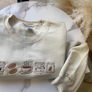 May include: Cream-colored sweatshirt with embroidered coffee-themed designs. The design includes a coffee cup, coffee beans, a cup of coffee, an espresso machine, and a bag of coffee beans. The sleeve is embroidered with the name "Jessica" and a heart.