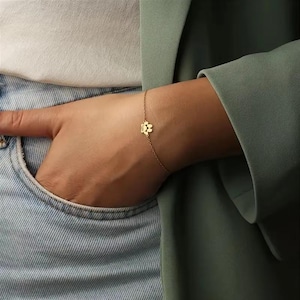 May include: A fine gold bracelet with a paw print pendant. The bracelet is displayed on a wrist, with a denim pocket and a green blazer in the background. The charm is a small, detailed paw print.