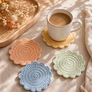 May include: A set of four round, crocheted coasters in pastel shades of peach, pale yellow, light blue, and mint green. Each coaster features a scalloped edge and a circular, textured stitch pattern. One yellow coaster supports a ceramic mug.