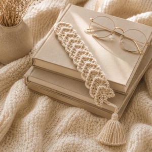 Peut inclure: Un marque-page en crochet de couleur crème composé d'une série de motifs en forme de cœur reliés entre eux et d'un pompon assorti à l'extrémité. Il est posé sur deux livres beiges empilés à côté d'une paire de lunettes rondes dorées.