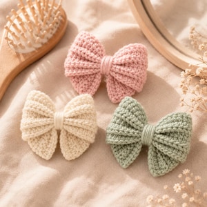 May include: Three crochet hair bows in soft pastel colors including light pink, cream, and sage green. Each bow features a ribbed texture with a central wrapped knot detail, crafted from yarn with a visible stitch pattern throughout the design.