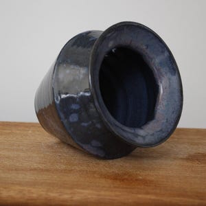 May include: A dark blue ceramic vase with a wide opening and a cylindrical body. The vase has a glossy finish and sits on a wooden surface. The interior of the vase is a darker shade of blue.