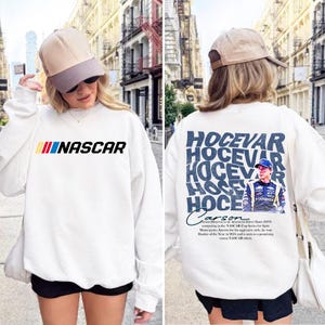May include: A white long-sleeve sweatshirt featuring the NASCAR logo on the front in black with yellow, red, and blue stripes. The back displays the name HOCEVAR in large blue text, a graphic of driver Carson Hocevar, and descriptive text.