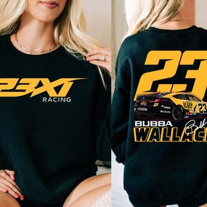 Könnte beinhalten: Schwarzes Sweatshirt mit dem Text "23XI Racing" in Gelb auf der Vorderseite. Auf der Rückseite sind die gelbe Zahl "23", eine gelb-schwarze Rennwagen-Grafik und der Name "Bubba Wallace" in Gelb abgebildet.