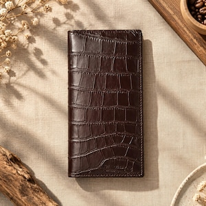 May include: A dark brown, rectangular wallet featuring a glossy, embossed crocodile skin texture. The wallet has a vertical orientation with visible stitching along the edges and a smooth, polished finish that reflects light across its surface.