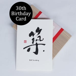 以下が含まれることがあります： 黒い書道と赤いスタンプが付いた白い30歳の誕生日カード。カードは赤いリボンが付いた木箱の上にあります。「Still building」というテキストがカードの下部にあります。